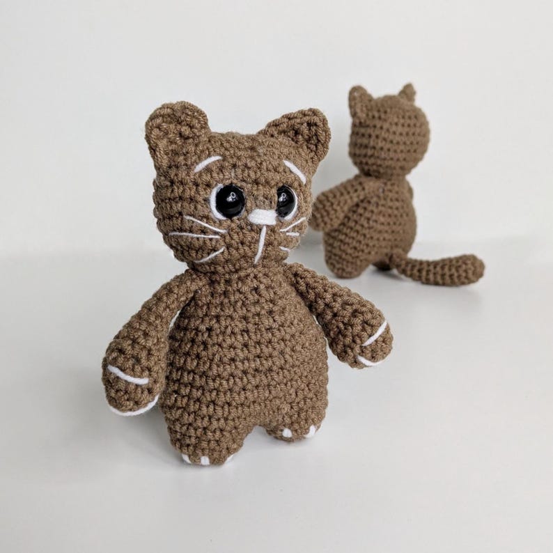 May include: Two brown crocheted cat figurines made with yarn. The front cat features black plastic eyes, white embroidered whiskers, and white accents on the paws. The second cat is positioned behind it, showing the back view with a small tail.