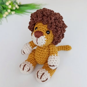May include: A crocheted lion toy with brown curly hair, a yellow body, and white paws. The lion is sitting with its tail curled behind it.