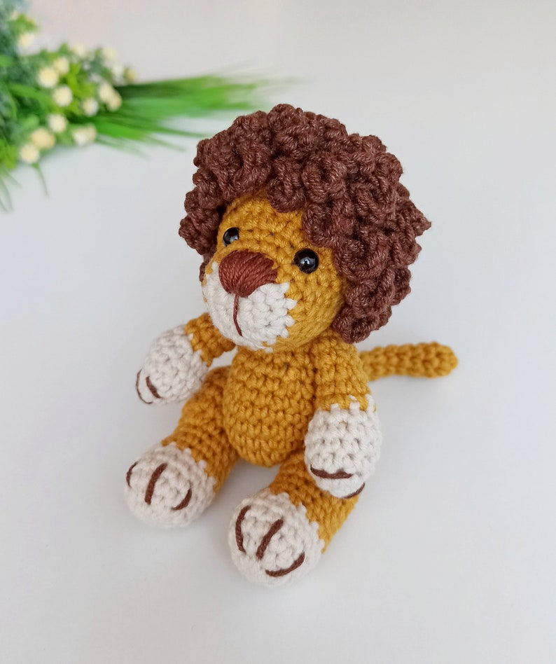 May include: A crocheted lion toy with brown curly hair, a yellow body, and white paws. The lion is sitting with its tail curled behind it.