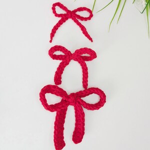 May include: Three handmade red crochet bows of varying sizes are arranged vertically against a white background. The bows are made of a thick, textured yarn, and each bow has a distinct shape. The top bow is the smallest, while the bottom bow is the largest.