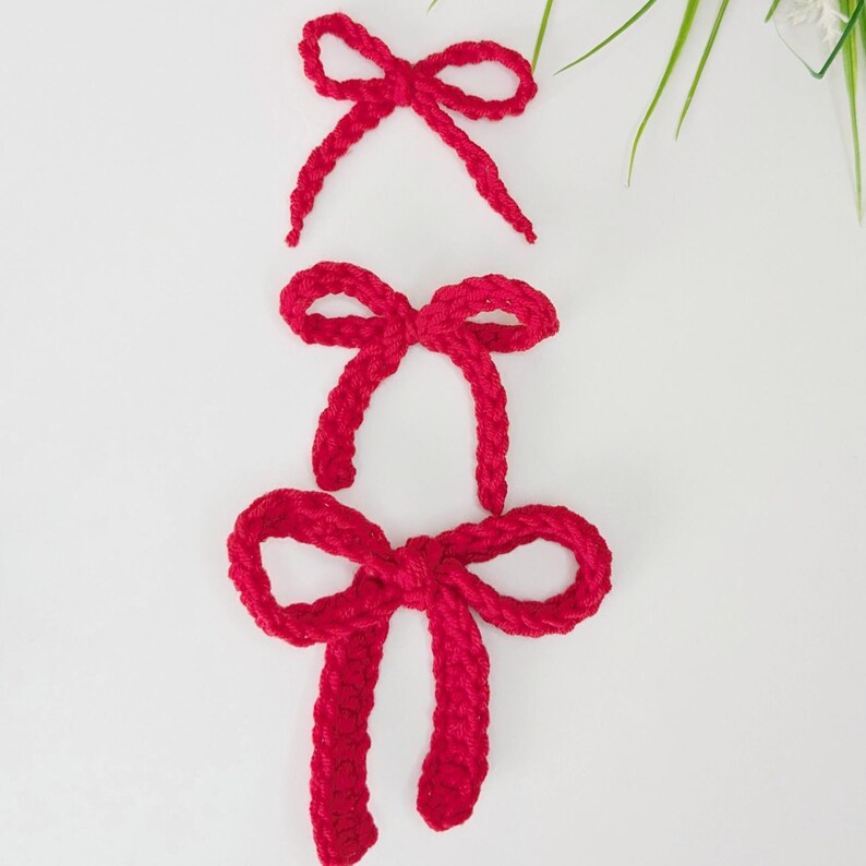 May include: Three handmade red crochet bows of varying sizes are arranged vertically against a white background. The bows are made of a thick, textured yarn, and each bow has a distinct shape. The top bow is the smallest, while the bottom bow is the largest.