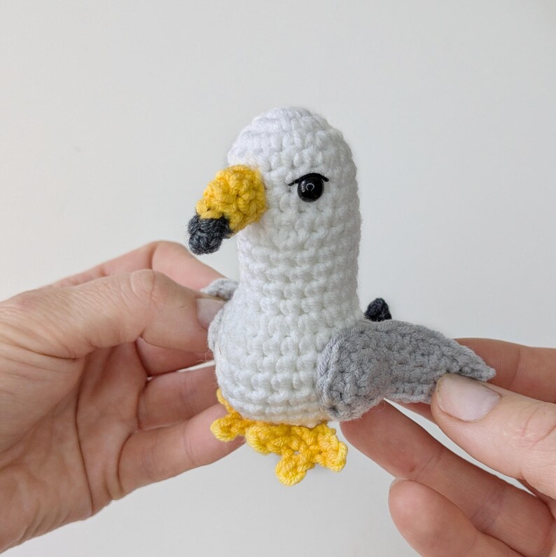 Op de afbeelding: Een klein, handgemaakt gehaakt meeuwspeeltje met een wit lichaam, lichtgrijze vleugels, een gele snavel met een donkergrijze punt en kleine gele pootjes. De vogel heeft &eacute;&eacute;n zwart kraaloog zichtbaar aan de zijkant van de kop, gemaakt van garen.