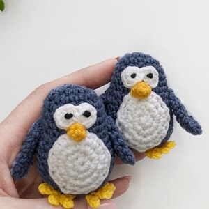 Pode incluir: Dois brinquedos de pinguim de croch&ecirc; com corpos azuis, barrigas brancas e p&eacute;s amarelos. Os pinguins s&atilde;o segurados em uma m&atilde;o.