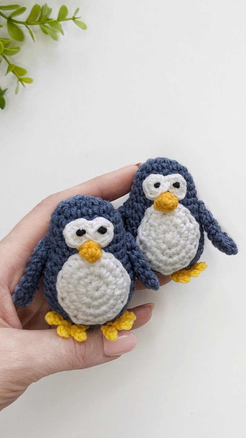 Pode incluir: Dois brinquedos de pinguim de croch&ecirc; com corpos azuis, barrigas brancas e p&eacute;s amarelos. Os pinguins s&atilde;o segurados em uma m&atilde;o.