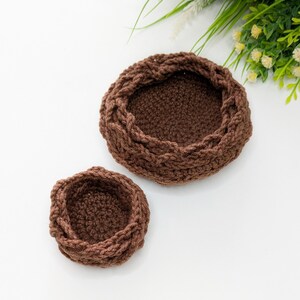 May include: Two round, dark brown crocheted bowls of different sizes. Each bowl features a thick, textured, braided-style rim and a dense, solid-stitch base. The yarn has a matte finish and a consistent deep brown colour throughout both items.