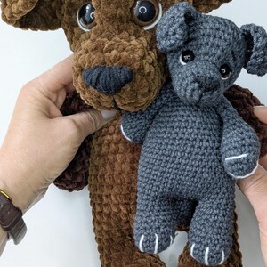 K&ouml;nnte beinhalten: Ein gro&szlig;er brauner Pl&uuml;schhund mit weicher, samtartiger Textur und dunkelbraunen Kunststoffaugen, der einen kleineren, dunkelgrauen H&auml;kelhund mit wei&szlig; gestickten Pfotendetails und kleinen schwarzen Kunststoffaugen h&auml;lt.