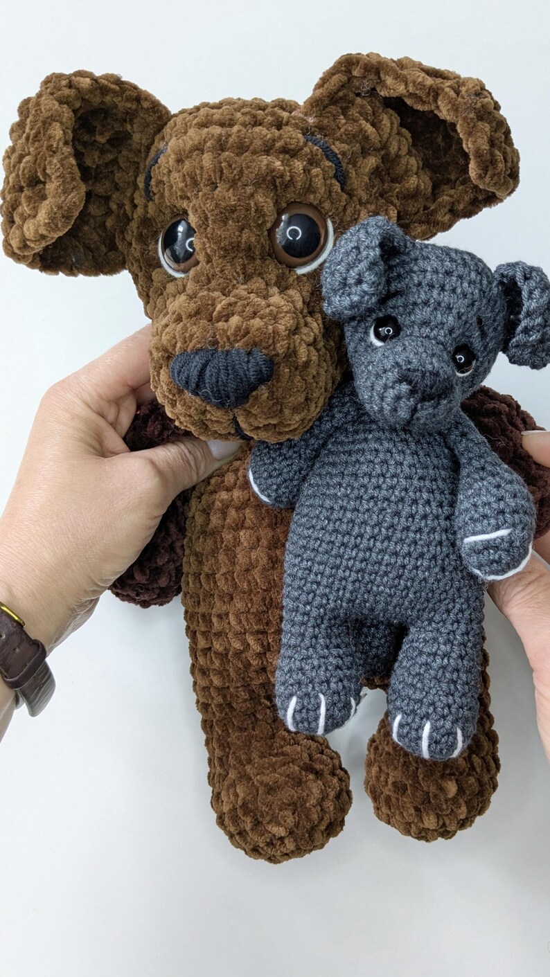 K&ouml;nnte beinhalten: Ein gro&szlig;er brauner Pl&uuml;schhund mit weicher, samtartiger Textur und dunkelbraunen Kunststoffaugen, der einen kleineren, dunkelgrauen H&auml;kelhund mit wei&szlig; gestickten Pfotendetails und kleinen schwarzen Kunststoffaugen h&auml;lt.