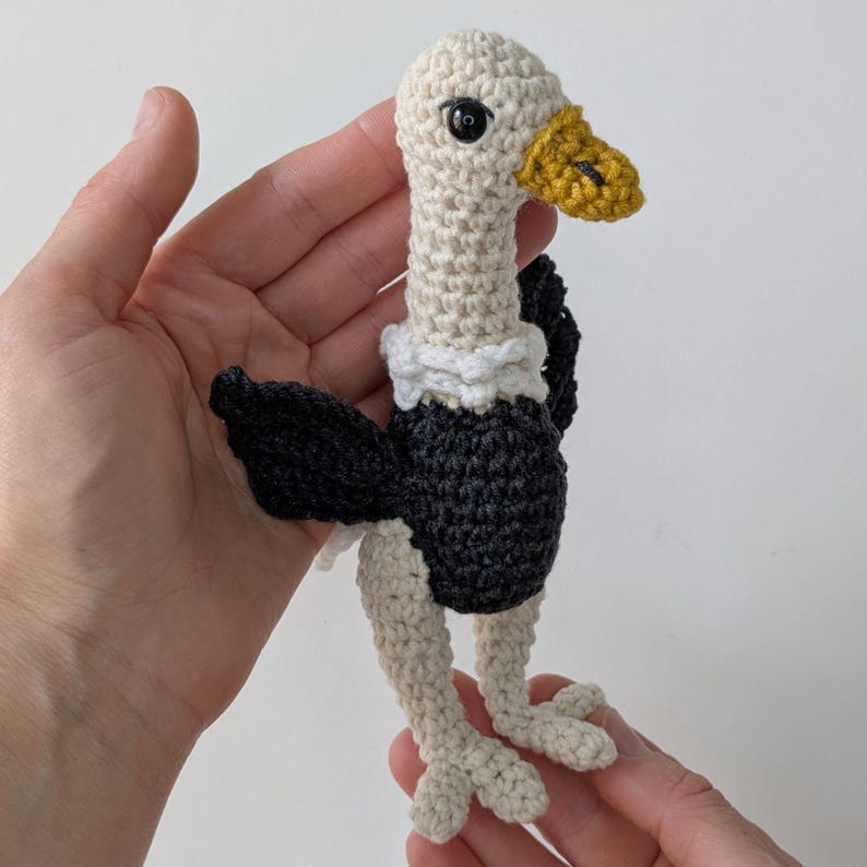 May include: A small crocheted vulture toy featuring a cream-colored head and neck, a white ruff, a dark charcoal body, and black wings. The bird has a textured yellow beak and black plastic safety eyes, crafted with visible yarn stitches.