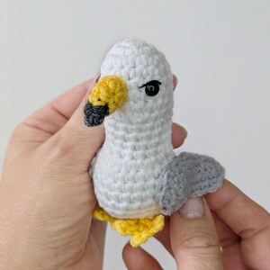 Op de afbeelding: Een klein, handgemaakt gehaakt meeuwfiguurtje met een witte kop en lichaam, grijze vleugels, een gele snavel met een zwarte punt, zwarte kraaloogjes en gele pootjes. De pluche vogel is gemaakt met zichtbare garensteken in een compacte vorm.