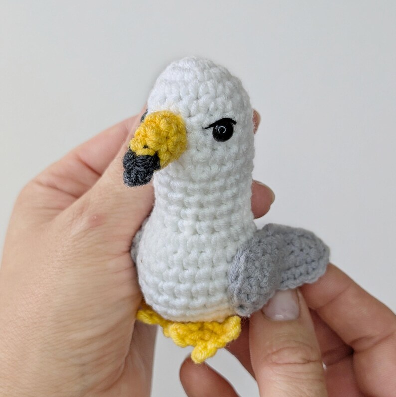 Op de afbeelding: Een klein, handgemaakt gehaakt meeuwfiguurtje met een witte kop en lichaam, grijze vleugels, een gele snavel met een zwarte punt, zwarte kraaloogjes en gele pootjes. De pluche vogel is gemaakt met zichtbare garensteken in een compacte vorm.