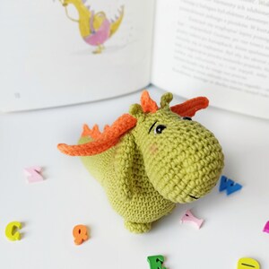 May include: A green crocheted dragon toy with orange wings and a cute face. The dragon is sitting on a white surface with colorful wooden letters scattered around it.