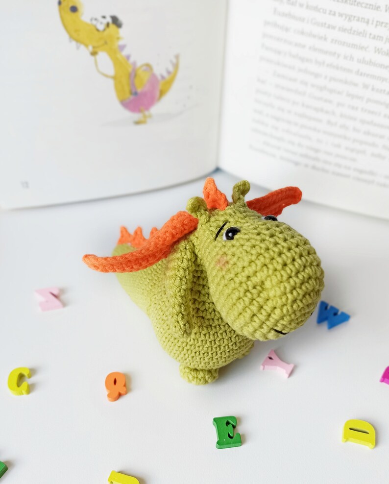 May include: A green crocheted dragon toy with orange wings and a cute face. The dragon is sitting on a white surface with colorful wooden letters scattered around it.