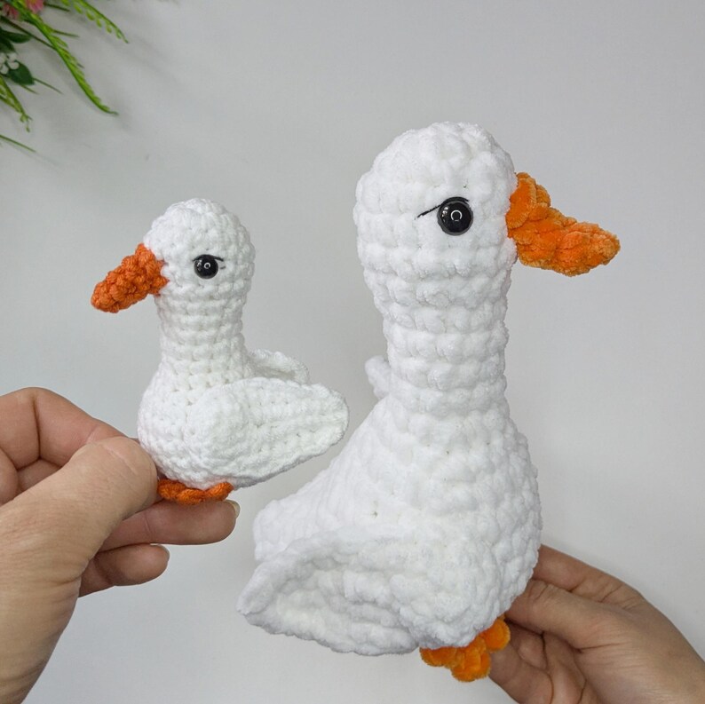 May include: Two handmade crochet plush toys shaped like geese. Each goose is crafted from white yarn with a textured, looped finish, featuring a bright orange beak and feet, and small black plastic bead eyes for a detailed, realistic appearance.