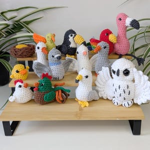 May include: A collection of small, handcrafted crochet bird figurines in various colors and species, including a pink flamingo, a white owl with spread wings, a toucan, a seagull, a rooster, and a nest with an egg, all arranged on a tiered wooden stand.