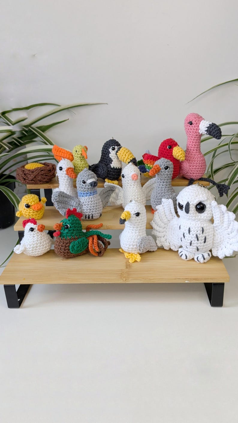 May include: A collection of small, handcrafted crochet bird figurines in various colors and species, including a pink flamingo, a white owl with spread wings, a toucan, a seagull, a rooster, and a nest with an egg, all arranged on a tiered wooden stand.