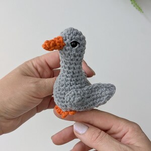 May include: A small, handmade crochet bird figurine crafted from grey yarn. The bird features a textured, knotted surface, a bright orange beak, and matching orange feet. It has a single visible black bead eye on the side of its head.
