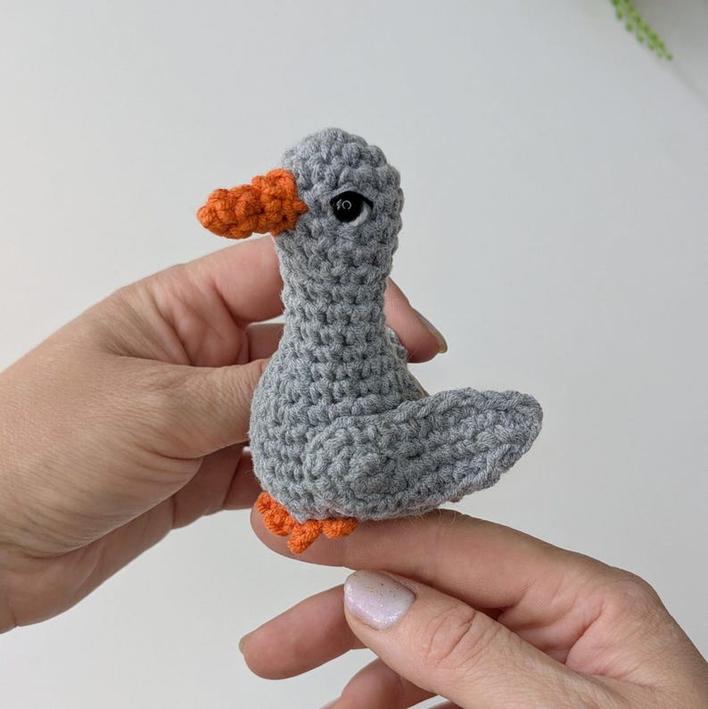May include: A small, handmade crochet bird figurine crafted from grey yarn. The bird features a textured, knotted surface, a bright orange beak, and matching orange feet. It has a single visible black bead eye on the side of its head.