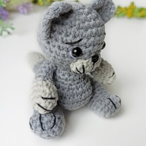 May include: A gray crocheted cat toy with black eyes and a black nose. The cat is sitting on a white surface.
