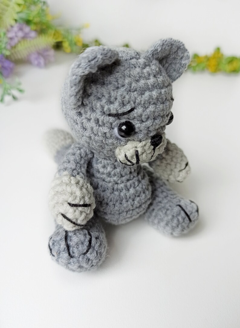 May include: A gray crocheted cat toy with black eyes and a black nose. The cat is sitting on a white surface.
