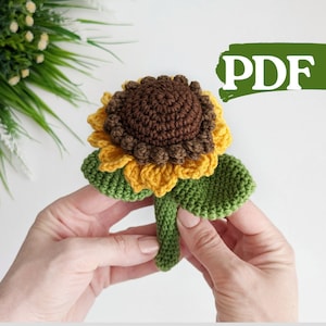 Puede incluir: Girasol de crochet con pétalos marrones y amarillos y hojas verdes. El girasol se sostiene en una mano. La imagen incluye la palabra "PDF".