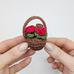 May include: A miniature crochet basket filled with red roses and green leaves. The basket is brown with a woven handle. The flowers are a vibrant red, and the leaves are a deep green. The basket is held in a person's hands.