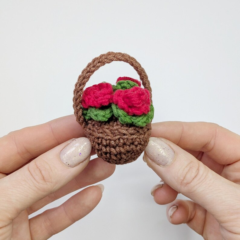 May include: A miniature crochet basket filled with red roses and green leaves. The basket is brown with a woven handle. The flowers are a vibrant red, and the leaves are a deep green. The basket is held in a person's hands.