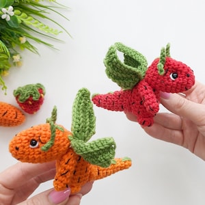 May include: Two crocheted dragon toys, one orange and one red, with green wings. The orange dragon is shaped like a carrot and the red dragon is shaped like a strawberry.