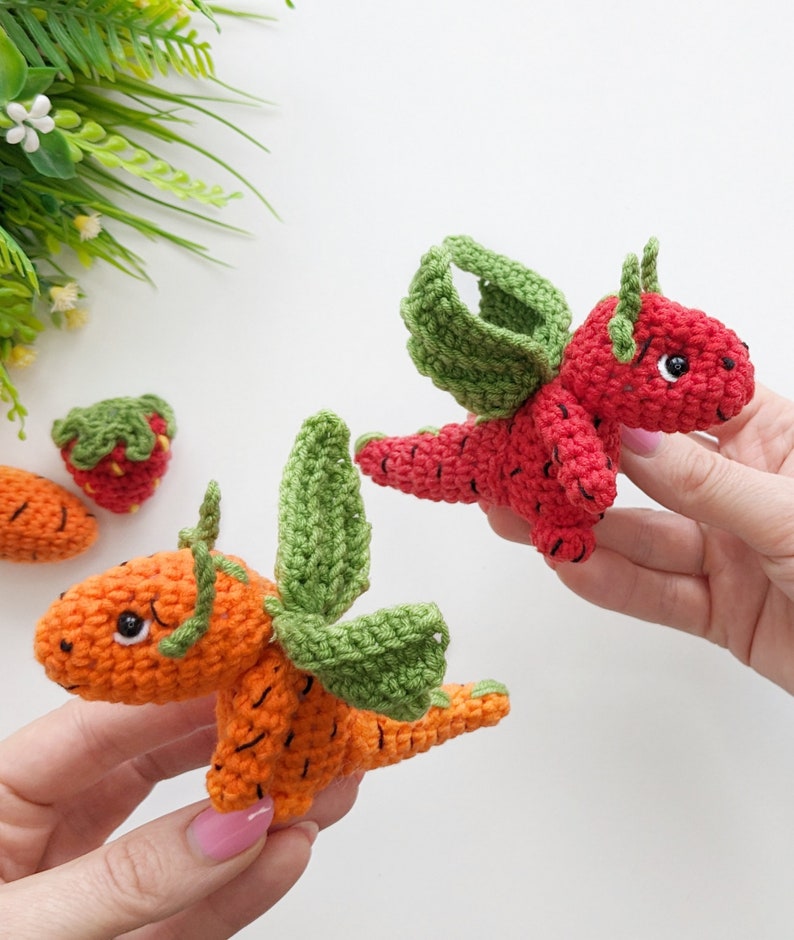 May include: Two crocheted dragon toys, one orange and one red, with green wings. The orange dragon is shaped like a carrot and the red dragon is shaped like a strawberry.