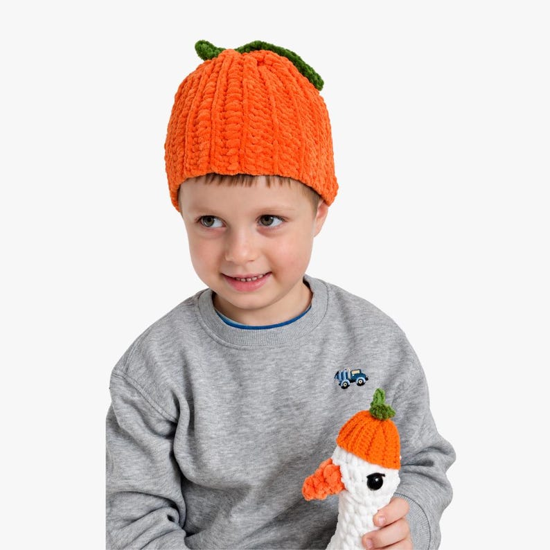 May include: A bright orange crocheted beanie hat with a textured, ribbed design and a small green leaf accent on the top. The hat is made from a soft, plush yarn material and features a rounded crown shape that fits snugly on the head.