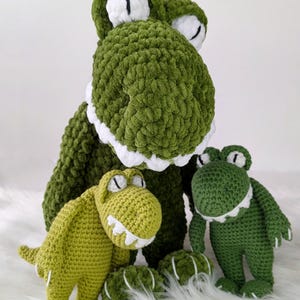 Puede incluir: Tres peluches de dinosaurio verdes tejidos a ganchillo con detalles blancos. El dinosaurio m&aacute;s grande est&aacute; en la parte trasera, con dos m&aacute;s peque&ntilde;os delante. Los juguetes tienen ojos y dientes blancos y est&aacute;n sobre una superficie blanca y esponjosa.