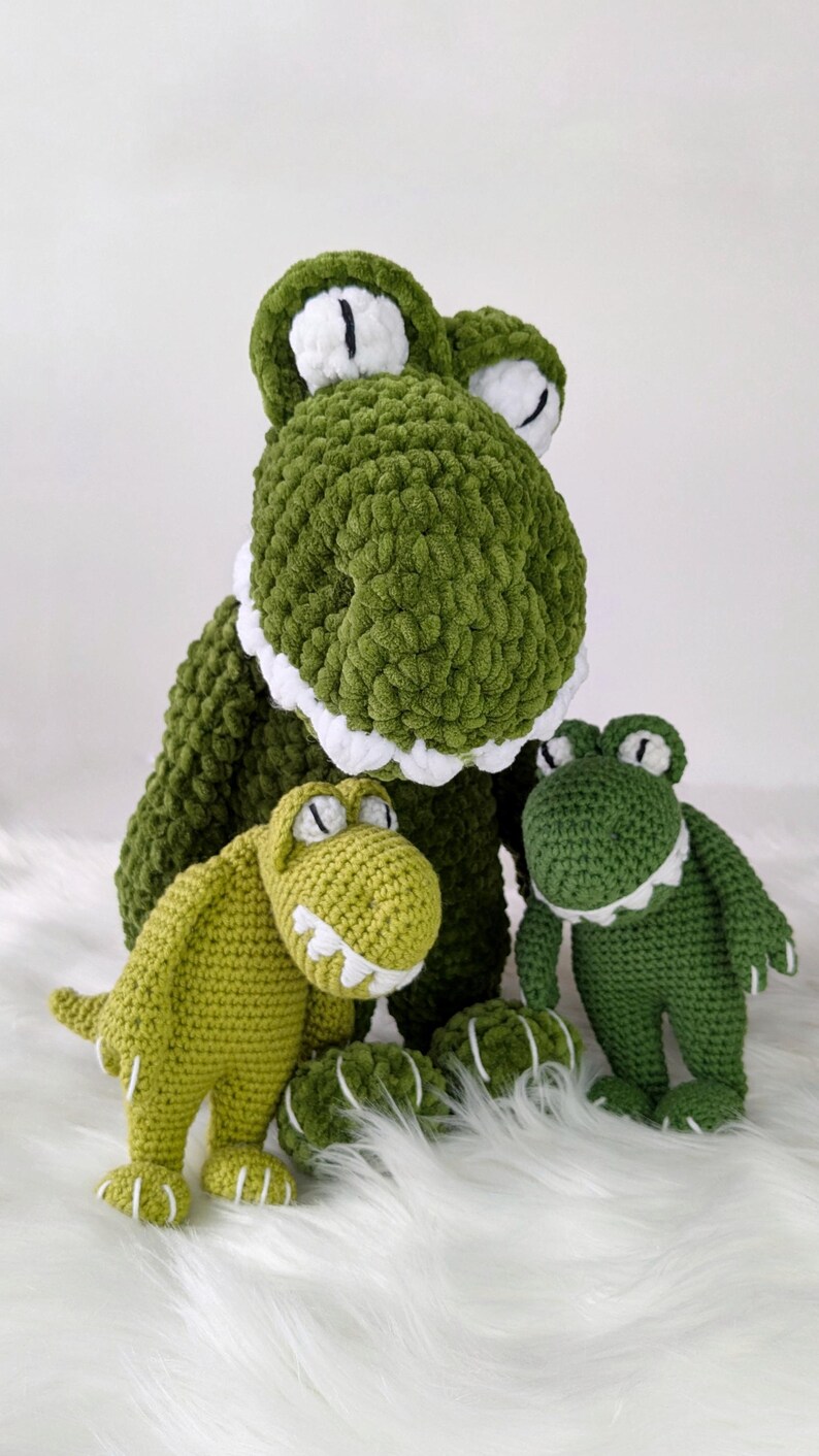 Puede incluir: Tres peluches de dinosaurio verdes tejidos a ganchillo con detalles blancos. El dinosaurio m&aacute;s grande est&aacute; en la parte trasera, con dos m&aacute;s peque&ntilde;os delante. Los juguetes tienen ojos y dientes blancos y est&aacute;n sobre una superficie blanca y esponjosa.