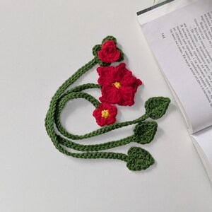 Puede incluir: Un marcap&aacute;ginas de ganchillo hecho a mano con tres flores rojas de centro amarillo y tallos verdes. Los tallos se extienden en largos cordones trenzados que terminan en peque&ntilde;as hojas verdes de ganchillo. El marcap&aacute;ginas est&aacute; junto a un libro.