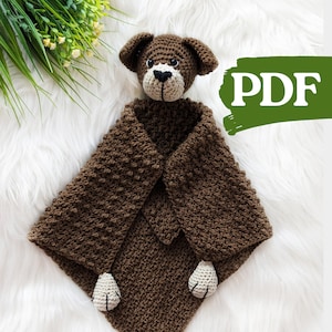Könnte beinhalten: Braune gehäkelte Babydecke mit einem angehängten gehäkelten Hunde-Kopf. Die Decke hat ein strukturiertes Muster und der Hund hat weiße Filzpfoten und schwarze Augen.