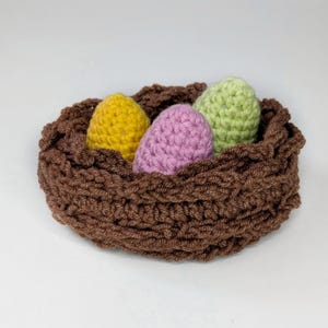May include: A brown crocheted nest containing three eggs in yellow, lilac, and green. The nest is made from thick, textured yarn, and the eggs appear to be made of a similar material. This handmade item is likely for Easter.