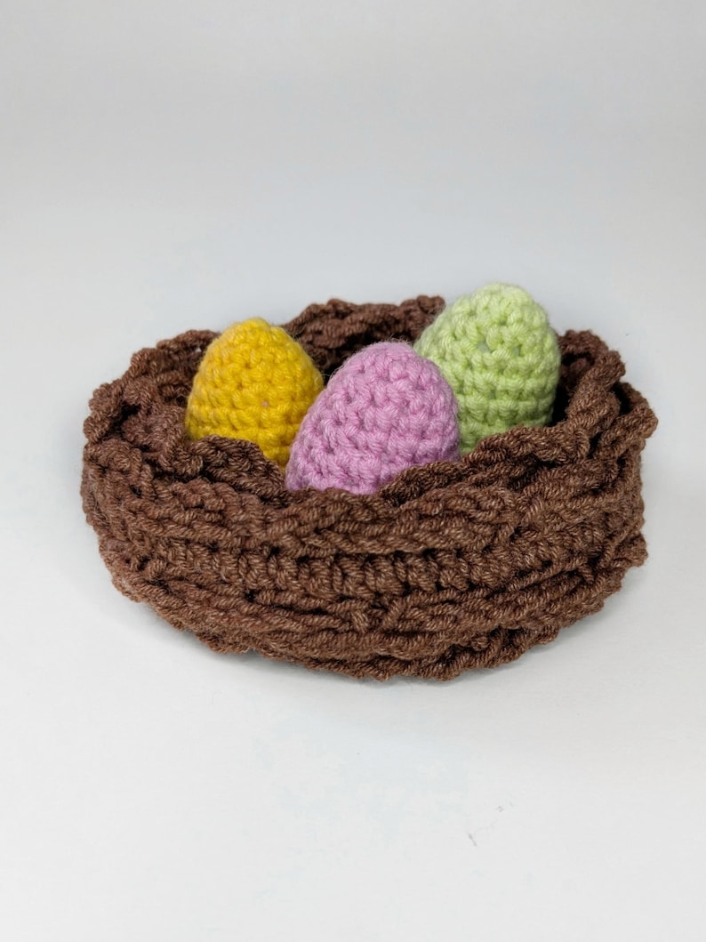 May include: A brown crocheted nest containing three eggs in yellow, lilac, and green. The nest is made from thick, textured yarn, and the eggs appear to be made of a similar material. This handmade item is likely for Easter.