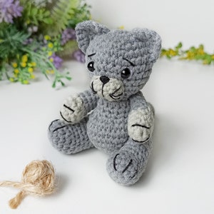 May include: A gray crocheted cat toy with black embroidered eyes and a black nose. The cat is sitting with its paws outstretched.