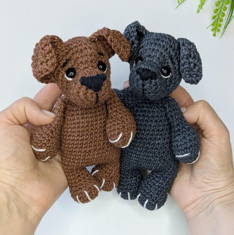 K&ouml;nnte beinhalten: Zwei kleine geh&auml;kelte Stofftiere in Hundeform. Ein Hund ist braun mit einer schwarzen Nase und wei&szlig; gestickten Pfotendetails, w&auml;hrend der andere dunkelgrau ist, ebenfalls mit einer schwarzen Nase und wei&szlig; gestickten Pfotendetails.