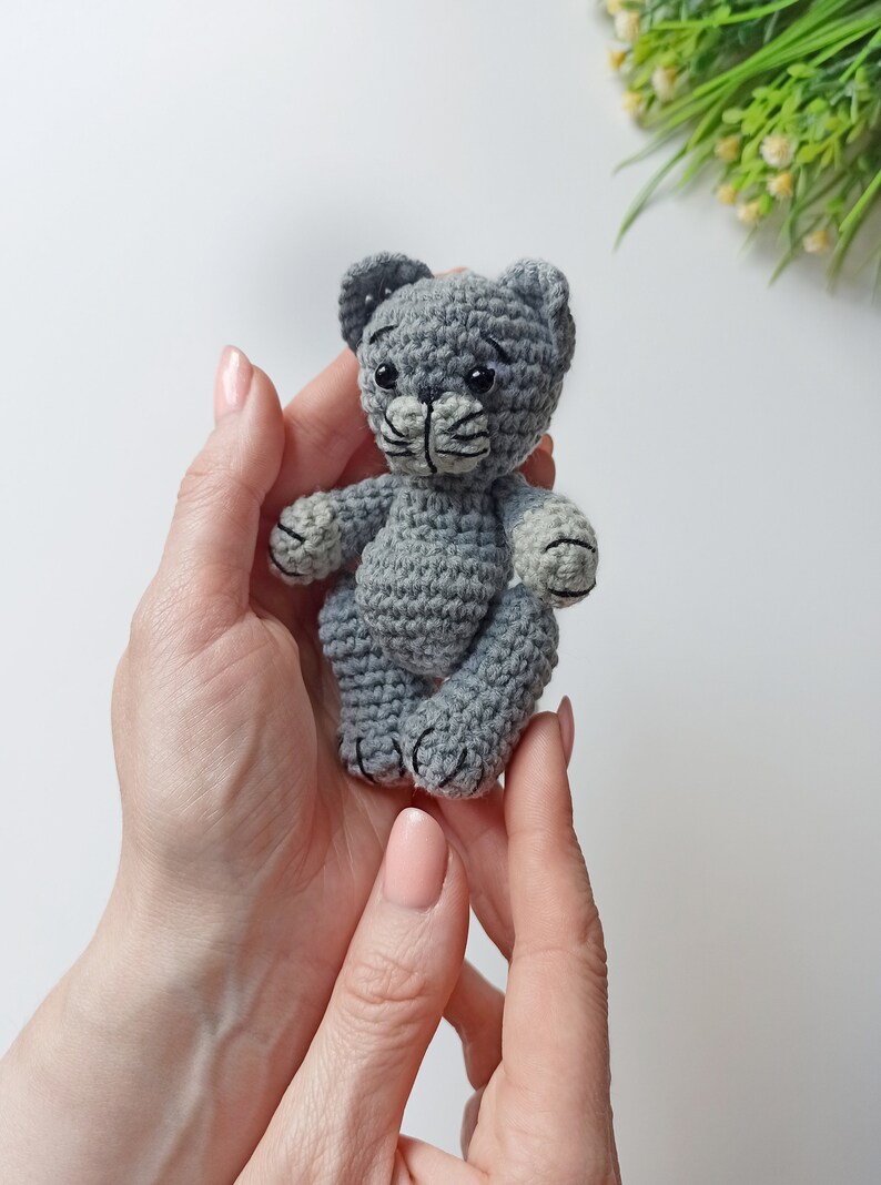 May include: A small gray crocheted cat toy with black eyes and a black nose. The cat is sitting in the palm of a person's hand.