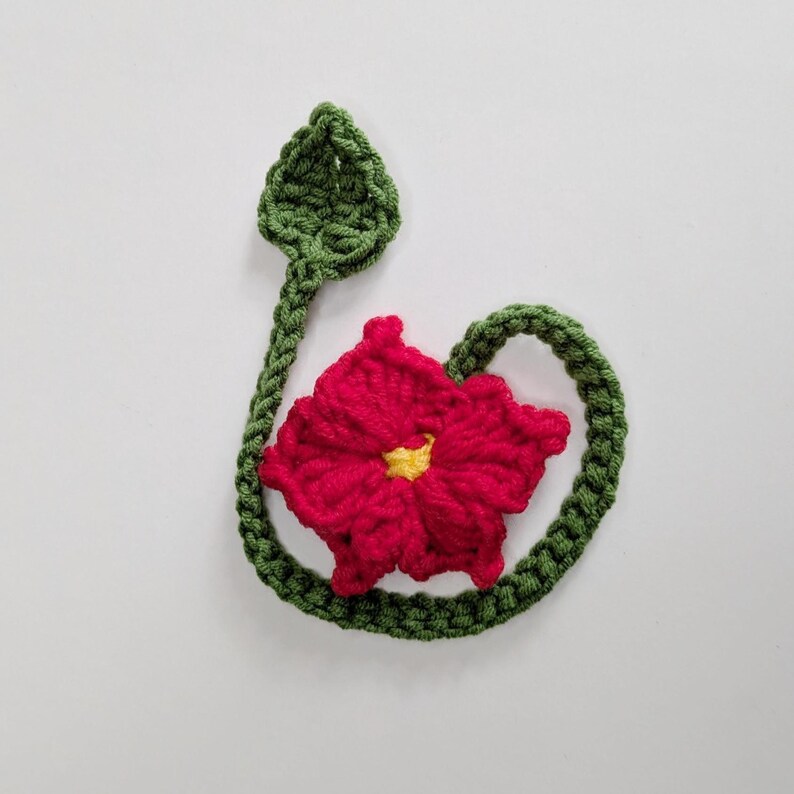 Puede incluir: Una aplicaci&oacute;n de flor tejida a ganchillo con una vibrante flor roja de cinco p&eacute;talos y un peque&ntilde;o centro amarillo. La flor est&aacute; unida a un tallo verde largo y sinuoso que se curva en un bucle y termina en una hoja verde puntiaguda.