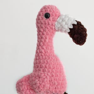 May include: A plush flamingo toy crafted from soft pink yarn with a textured, looped finish. The bird features a white and dark brown beak, a single visible black plastic eye, and long, dangling dark brown legs made of the same plush material.