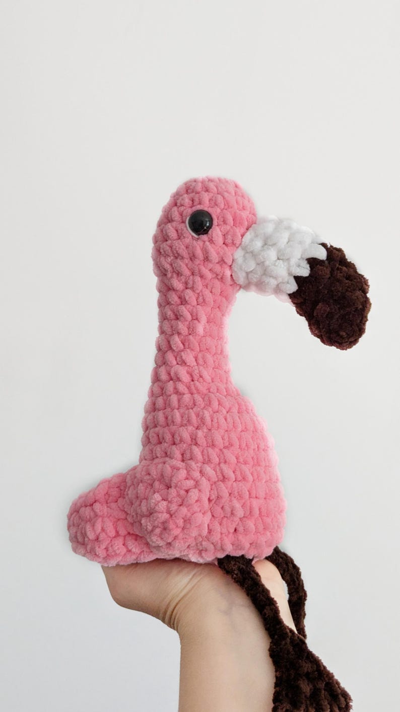 May include: A plush flamingo toy crafted from soft pink yarn with a textured, looped finish. The bird features a white and dark brown beak, a single visible black plastic eye, and long, dangling dark brown legs made of the same plush material.