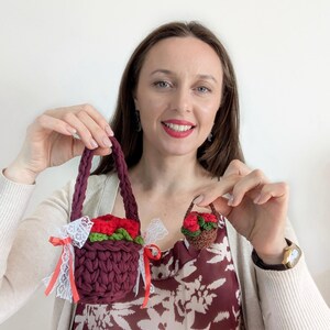 May include: Two handmade crochet baskets are displayed. The larger basket is burgundy with red roses and green leaves, accented with white lace and red ribbons. The smaller basket is brown with red roses. Both have braided handles.