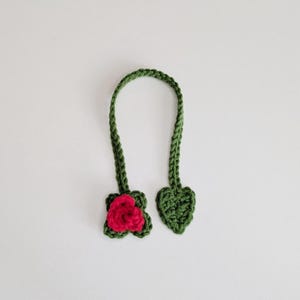 Puede incluir: Un marcap&aacute;ginas de ganchillo verde con una larga tira trenzada que conecta una rosa de color rosa vibrante en un extremo y una hoja en forma de coraz&oacute;n en el otro. La textura del hilo es visible en todo el accesorio hecho a mano.