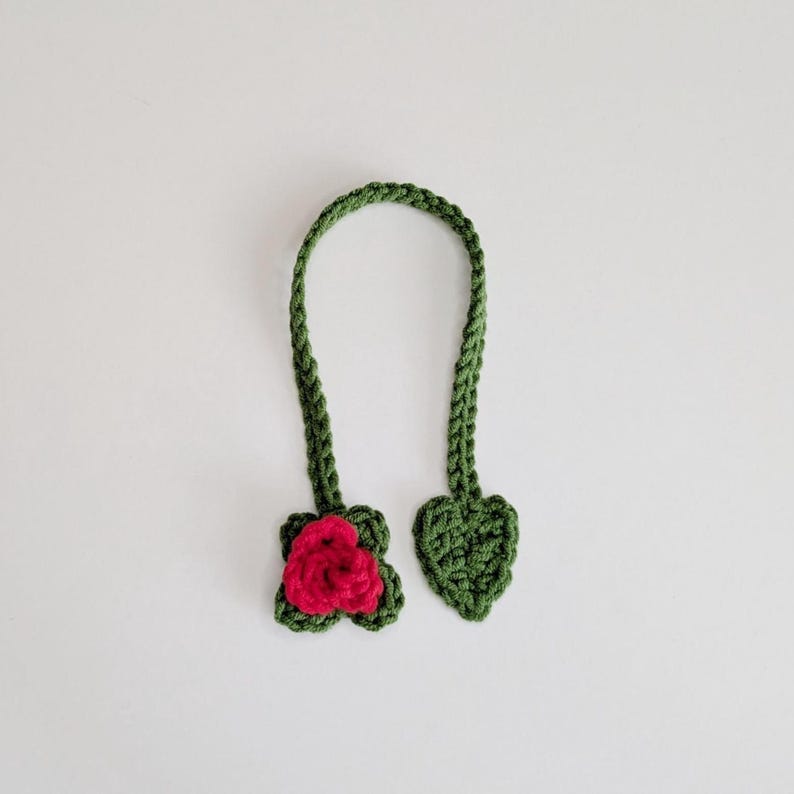 Puede incluir: Un marcap&aacute;ginas de ganchillo verde con una larga tira trenzada que conecta una rosa de color rosa vibrante en un extremo y una hoja en forma de coraz&oacute;n en el otro. La textura del hilo es visible en todo el accesorio hecho a mano.