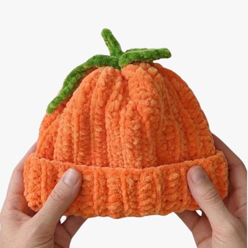 May include: A bright orange beanie made of soft, plush yarn with a ribbed texture. The hat features a green stem and leaf detail attached to the top center, mimicking the appearance of a pumpkin. The base of the beanie has a folded cuff.