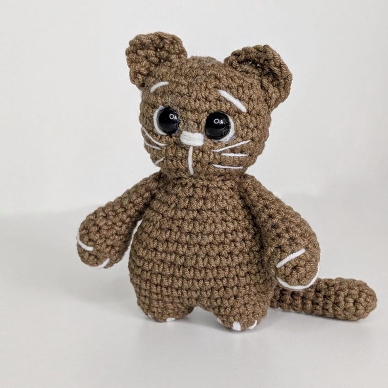 May include: A brown crocheted cat figurine featuring large black plastic eyes, white embroidered eyebrows, white whiskers, and white stitched details on the paws and tail. The stuffed animal has a rounded body, pointed ears, and a long tail.