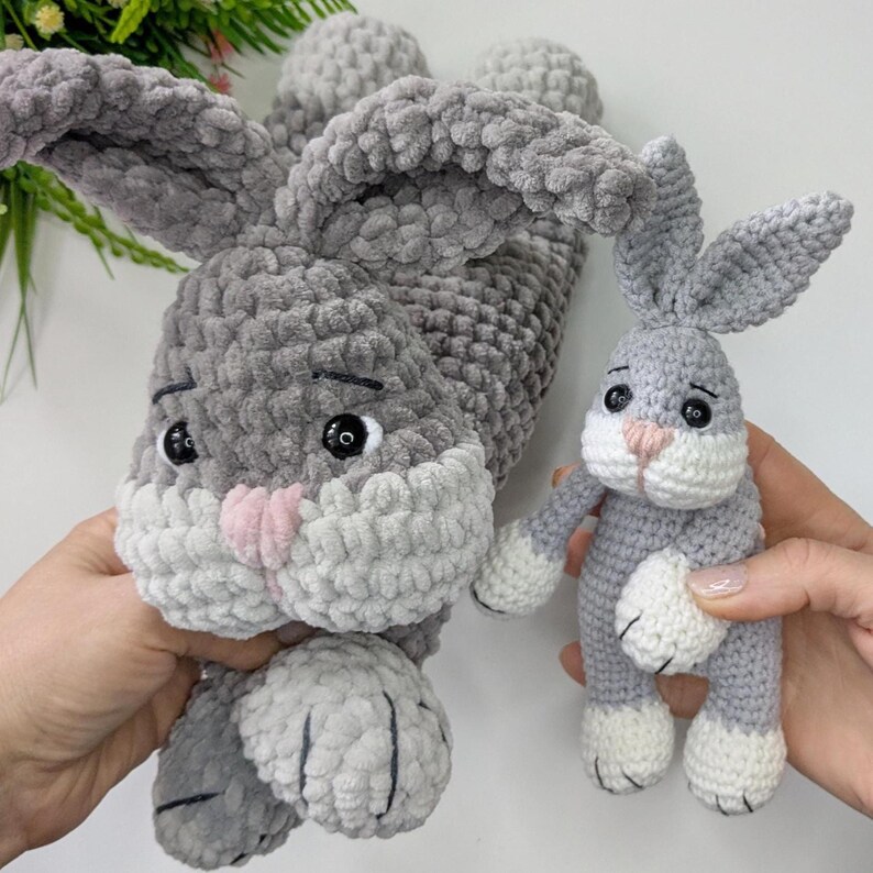 Op de afbeelding: Twee grijze en witte gehaakte konijnen. E&eacute;n groot konijn ligt en een kleiner konijn staat rechtop. Beide hebben roze neuzen, zwarte ogen en witte poten. De konijnen zijn handgemaakt en hebben een zachte, pluche textuur.
