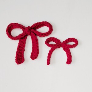May include: Two red crocheted bows of different sizes are displayed against a white background. The larger bow has a more defined knot and longer tails, while the smaller bow is simpler in design. The bows are made of a textured yarn.