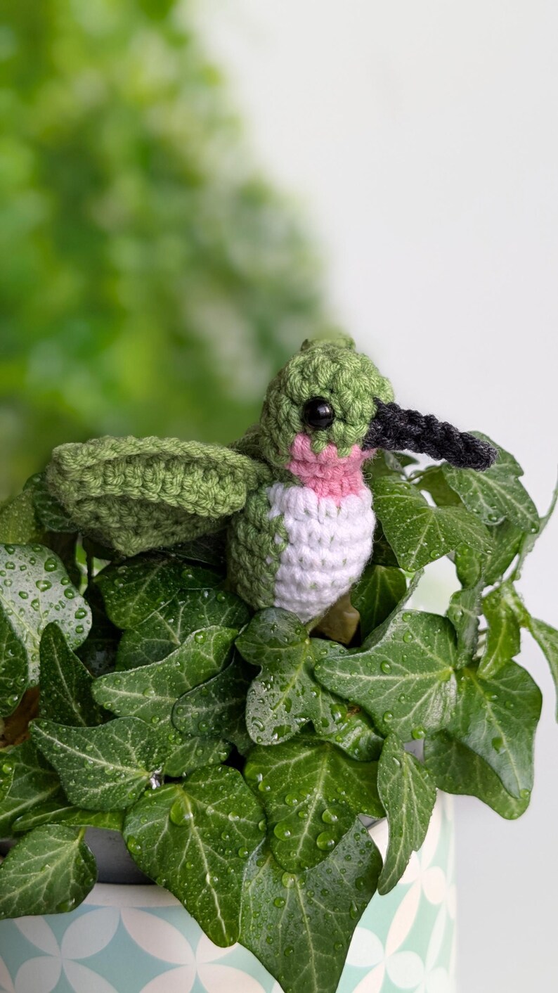 May include: A crocheted hummingbird with green, white and pink accents perched on a green leafy plant. The hummingbird has black eyes and a black beak.