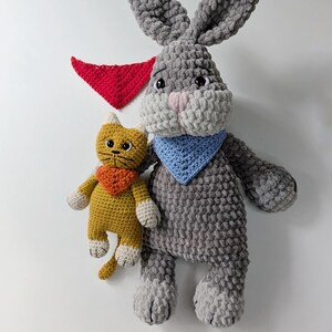 May include: A large gray crocheted rabbit with long ears and a light blue bandana, a smaller yellow crocheted cat with an orange bandana, and a separate red triangular bandana. The rabbit has a pink nose and black eyes, while the cat has black eyes.